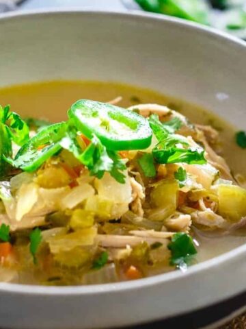 A bowl of chicken chili topped with sliced jalapeños, cilantro, and a lime wedge, served on a woven placemat with a spoon beside it.