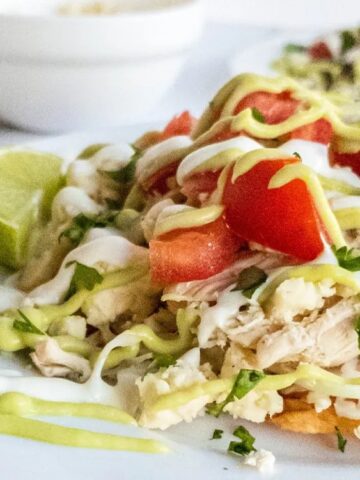 Two tostadas topped with shredded chicken, chopped tomatoes, cheese, cilantro, and drizzled with sour cream and avocado sauce, served with a lime wedge on a white plate.