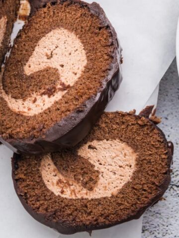 Two slices of chocolate Swiss roll cake with a cream filling are shown on parchment paper, next to a white plate with several metal forks.