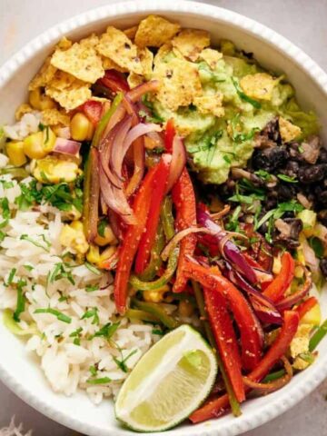 A bowl containing rice, black beans, sautéed red onions and peppers, guacamole, corn, crushed tortilla chips, and a lime wedge.