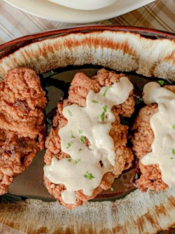 An oval plate with three pieces of fried steak, two topped with white gravy and herbs, sits on a plaid cloth.