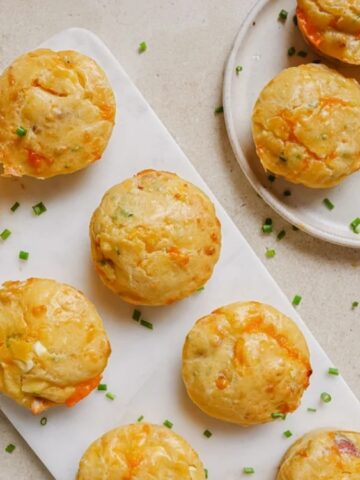 Nine savory muffins with bits of cheese and herbs are arranged on a white board and a round plate, topped with chopped chives on a light surface.