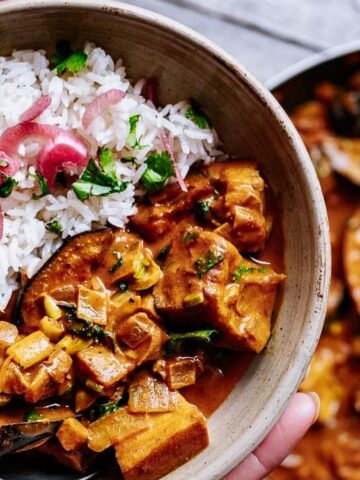A bowl of white rice topped with pickled onions and cilantro, served with a vegetable curry in a creamy sauce; a pan of curry and wooden spoon are in the background.