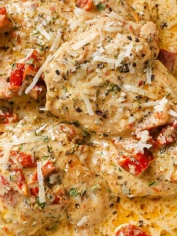 Chicken breasts in a creamy sauce with sun-dried tomatoes and herbs, topped with grated cheese, served on a light-colored plate.