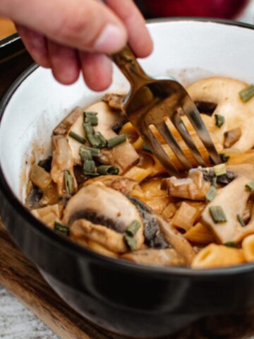 A hand holds a fork over a bowl of pasta with mushrooms and chopped herbs in a creamy sauce, placed on a wooden board.