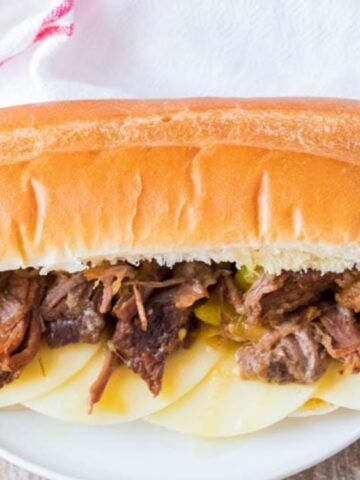 A sandwich roll filled with shredded beef, sliced cheese, and yellow-green peppers on a white plate.
