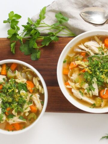 Two bowls of chicken noodle soup with carrots, garnished with fresh herbs, placed on a wooden board with cutlery, parsley, and dill nearby.