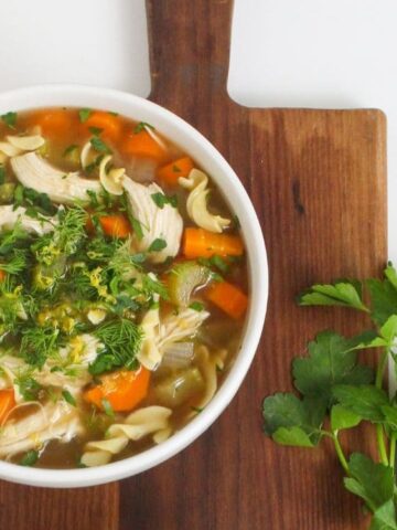 A bowl of chicken noodle soup with carrots and herbs sits on a wooden cutting board next to fresh parsley.
