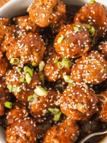 A bowl of glazed meatballs topped with sesame seeds and chopped green onions, placed on a wooden surface.
