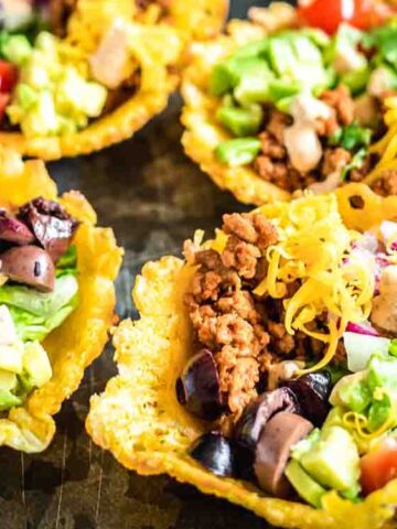 Four taco bowls made of melted cheese are filled with ground meat, chopped tomatoes, lettuce, avocado, red onions, olives, and shredded cheese, arranged on a dark plate.