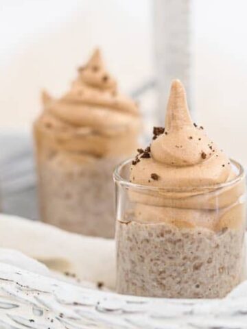 Three small glass cups filled with a creamy, beige dessert are arranged in a white wicker basket, topped with a swirl of mousse and a sprinkle of chocolate crumbs.