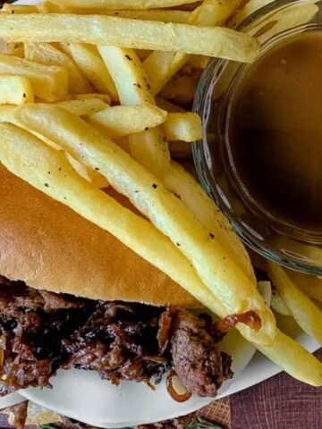 A plate with a beef sandwich and French fries, served with a glass bowl of brown gravy for dipping.