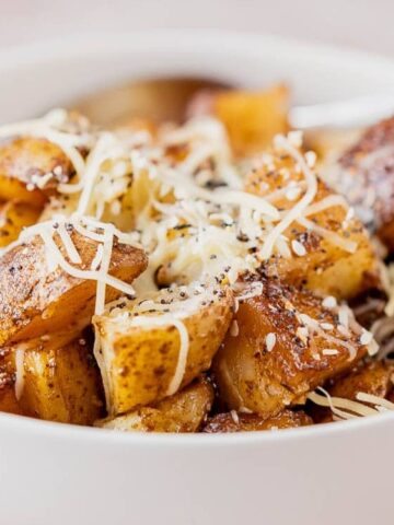 A white bowl filled with seasoned roasted potato cubes topped with shredded cheese, with a spoon in the bowl.