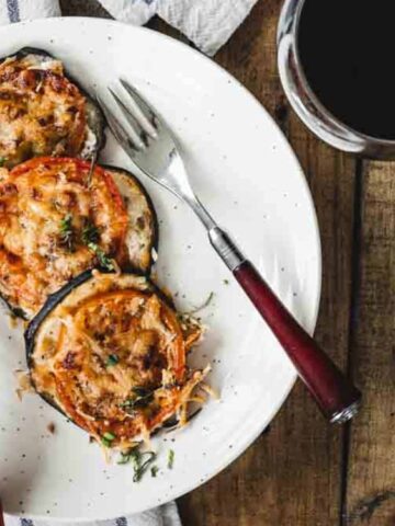 A plate with two baked eggplant slices topped with melted cheese and herbs, served with a knife and fork, next to a glass of red wine on a wooden table.