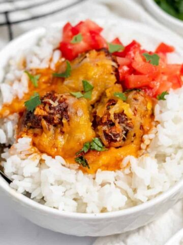 A bowl of white rice topped with cheesy meatballs, diced tomatoes, and chopped cilantro, with a spoon on the side.