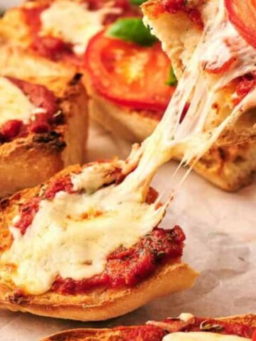 Slices of French bread pizza topped with melted cheese, tomato sauce, and tomato slices, with cheese stretching as one slice is lifted.