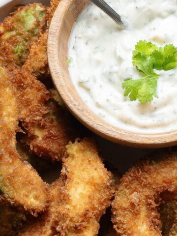 A plate of breaded and fried avocado slices served with a bowl of creamy dip garnished with cilantro.