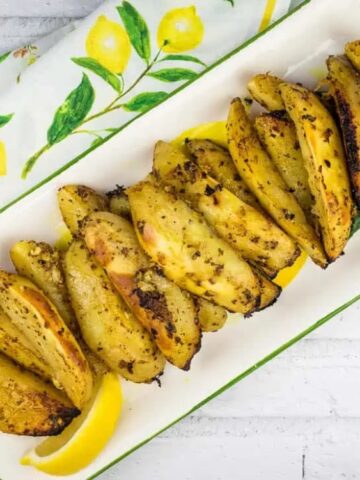 A rectangular plate with baked potato wedges garnished with herbs and spices, arranged in a row, with lemon wedges on the side.