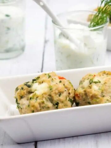 Two vegetable and herb dumplings served in a white dish, with a glass of creamy dipping sauce and a garnish of dill in the background.