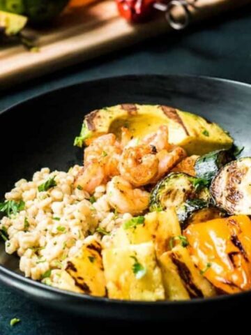 A black bowl containing grilled shrimp, zucchini, bell peppers, avocado, and barley, garnished with herbs. A fork and a cutting board with more vegetables are nearby.