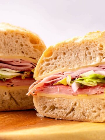 A sub sandwich cut in half, filled with sliced meats, cheese, lettuce, and onions, on a wooden board.