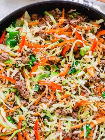 A skillet filled with cooked ground meat, shredded cabbage, sliced carrots, red bell pepper, and green onions on a striped cloth.