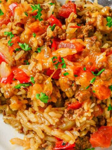 A plate of rice casserole with ground meat, red bell peppers, melted cheese, chopped onions, and sprinkled fresh parsley, served with a spoon.