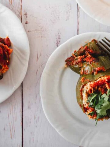 Three plates with stuffed bell peppers filled with a tomato-based mixture and garnished with herbs, each plate including a fork, on a white wooden surface.