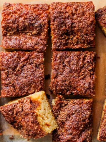 Twelve pieces of moist, golden-brown coffee cake with a crumbly cinnamon topping are arranged on a piece of parchment paper.