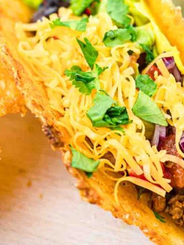 A close-up of a taco with a crispy shell filled with seasoned ground beef, shredded cheese, chopped lettuce, and fresh cilantro.