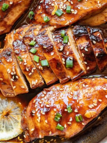 Sliced glazed chicken breast in a brown sauce, garnished with chopped chives and sesame seeds, with lemon slices in the pan.