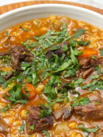 A bowl of beef and vegetable soup topped with chopped fresh herbs, placed on a wooden surface next to a spoon.