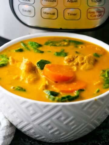 A white bowl filled with yellow curry containing spinach, carrots, and chicken sits in front of an electric pressure cooker.