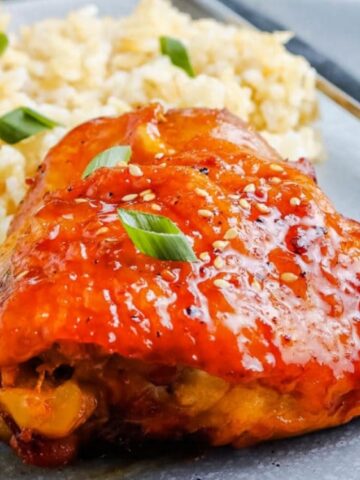 A glazed baked chicken thigh garnished with green onions is served alongside a portion of rice on a gray plate with a fork.