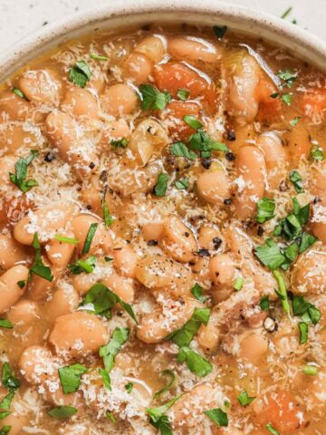 A bowl of bean soup garnished with chopped parsley and grated cheese, with a spoon resting inside.