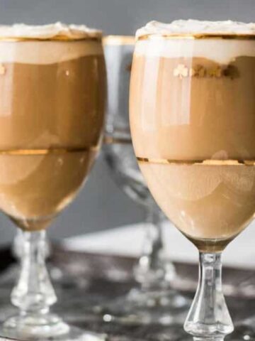 Four glass goblets, two filled with creamy layered coffee topped with foam and two empty, are arranged on a metallic tray.