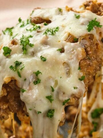 A close-up of a cheesy casserole being lifted from a skillet, topped with melted cheese and chopped parsley.