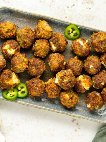 A rectangular plate holds several cooked meatballs garnished with sliced jalapeños. A small bowl of dipping sauce and shredded cheese are nearby on the table.