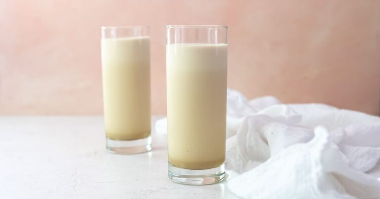 Two tall, clear glasses filled with a light beige liquid are placed on a white surface with a white cloth beside them and a pale pink background.