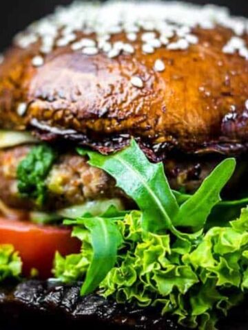 A burger with leafy lettuce, tomato, and greens, served in a bun made from two large portobello mushrooms topped with sesame seeds.