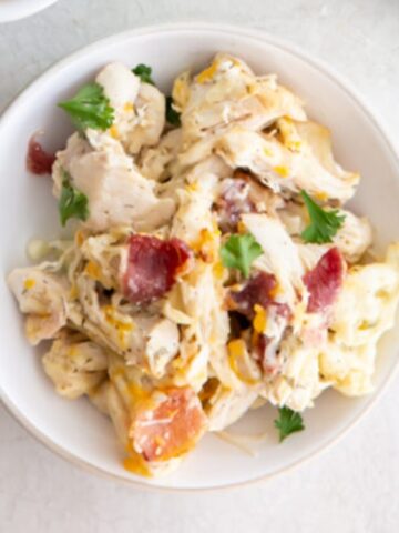 A white bowl filled with shredded chicken, cheese, chopped bacon, and fresh parsley on a light-colored surface.