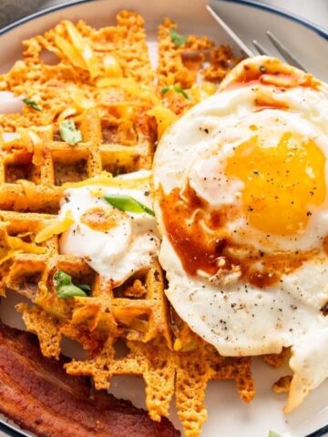 A plate with a fried egg, two strips of bacon, and a waffle topped with melted cheese and green onions.