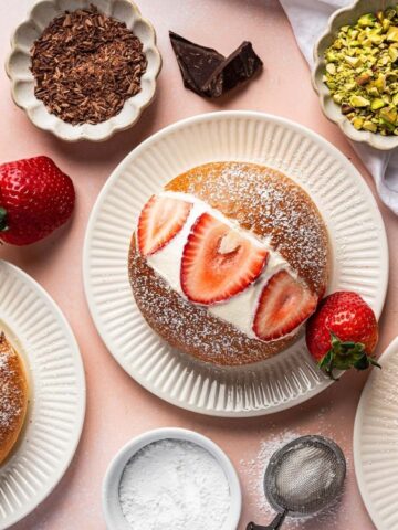 Several cream-filled pastries are displayed on white plates, topped with strawberries, pistachios, or chocolate shavings, surrounded by ingredients and a dusting of powdered sugar.