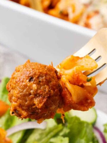 A fork holds a meatball and pasta with tomato sauce, with a salad and a casserole dish in the background.