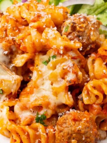 A plate of rotini pasta with tomato sauce, melted cheese, and meatballs, garnished with herbs, served with a side salad.