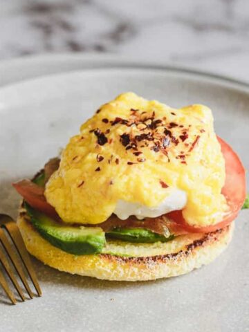 An open-faced breakfast sandwich with avocado, tomato, smoked salmon, poached egg, and scrambled eggs on an English muffin, served on a plate with a fork.
