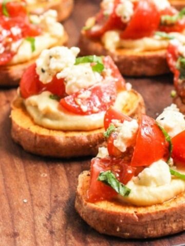 Slices of roasted sweet potato topped with hummus, cherry tomatoes, crumbled cheese, and herbs are arranged on a wooden board.
