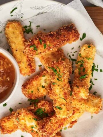 A plate of breaded, fried mozzarella sticks garnished with chopped herbs, served with a bowl of dipping sauce on the side.