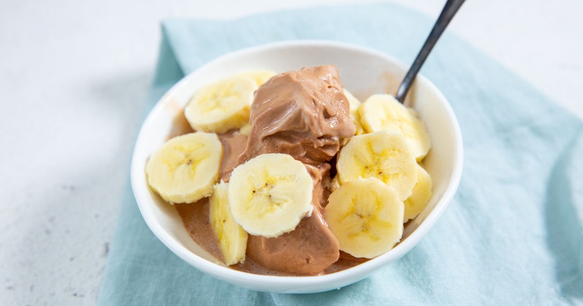 A bowl of chocolate ice cream topped with banana slices, with a spoon resting in the bowl, placed on a light blue cloth.