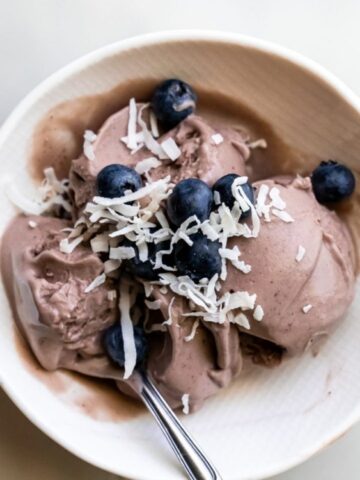 A bowl of chocolate ice cream topped with shredded coconut and fresh blueberries, with a spoon placed inside.
